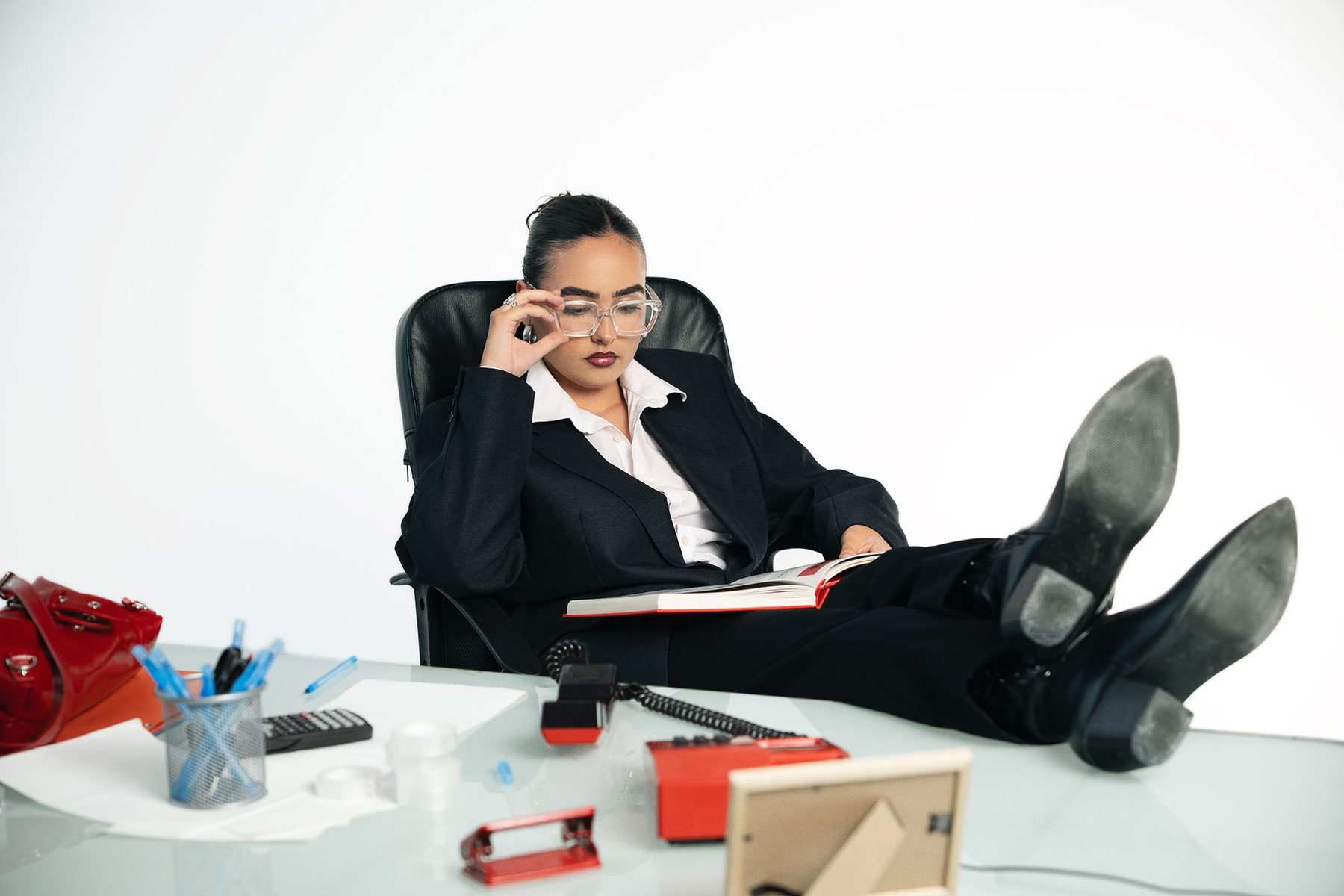 Aida in a black suit reading at a desk with her boots up, red telephone and red bag in the foreground