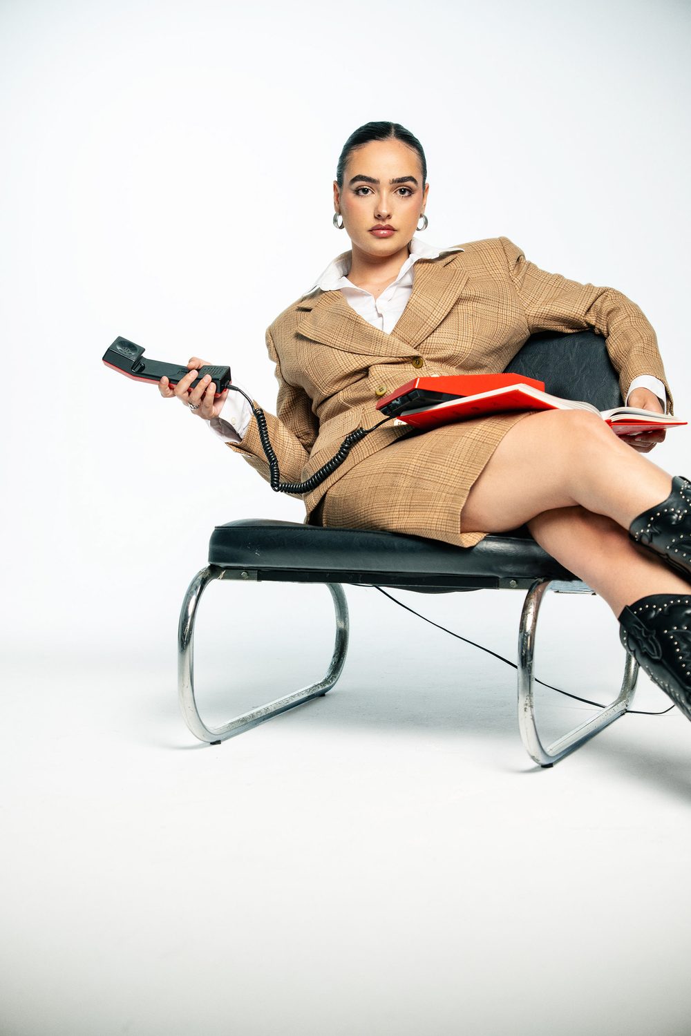 Aida seated in a chrome chair holding a red telephone in a camel checked suit and black boots