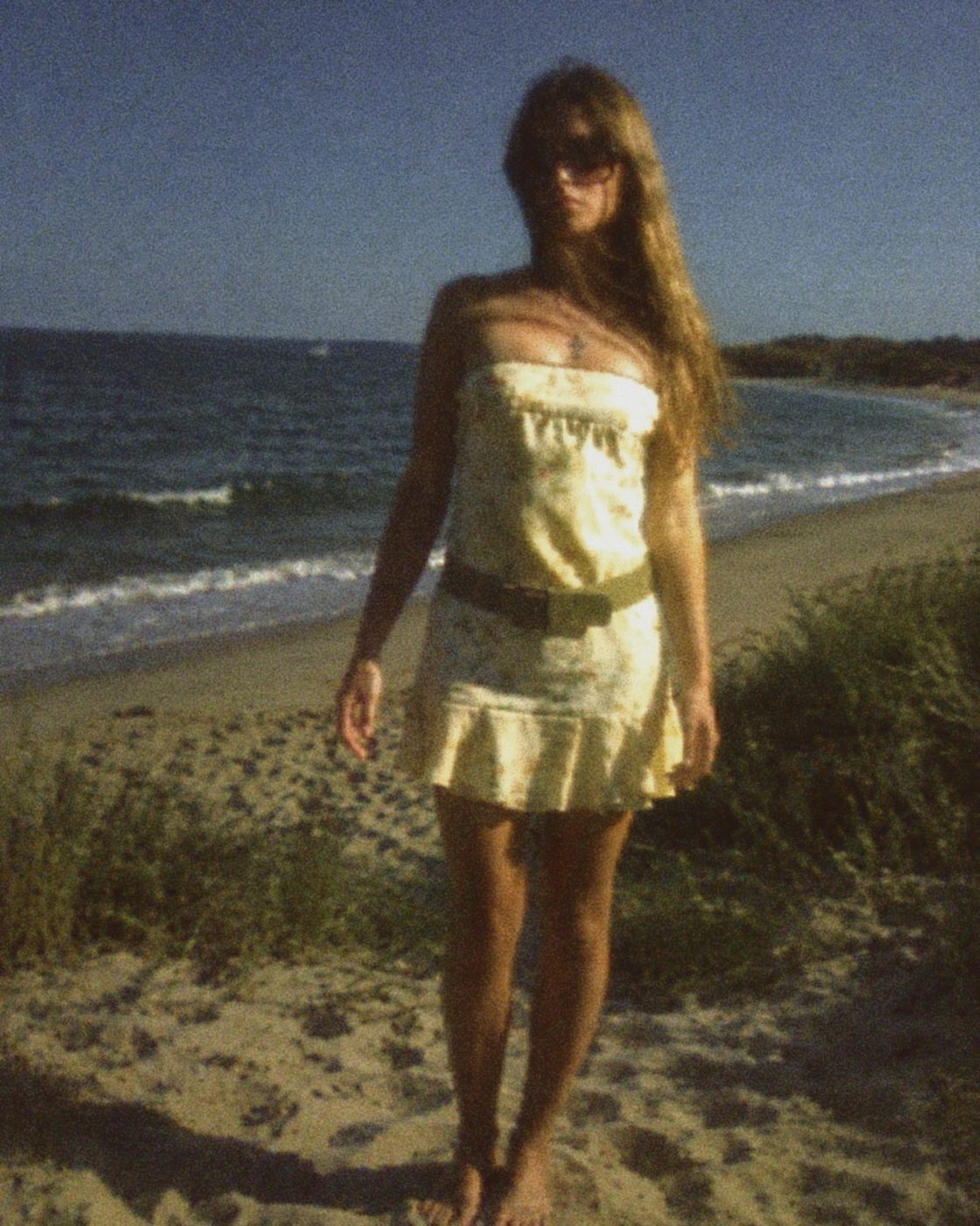 Model on a beach at dusk in a pale yellow ruffled strapless dress, sunglasses and a belt