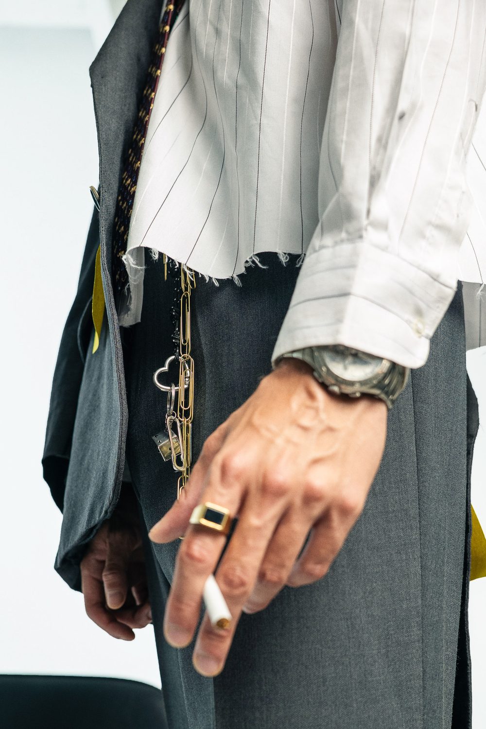 Detail of a raw hem pinstripe shirt under a grey blazer, a watch, a signet ring and a paper clip chain