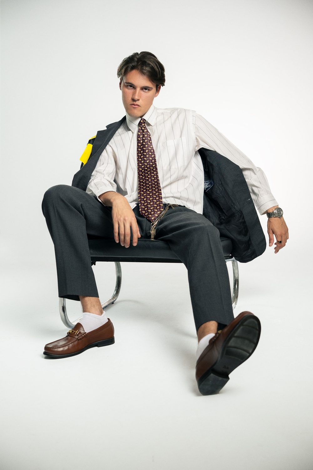 Marc seated in a chrome chair in a grey suit, striped shirt and brown loafers with white socks
